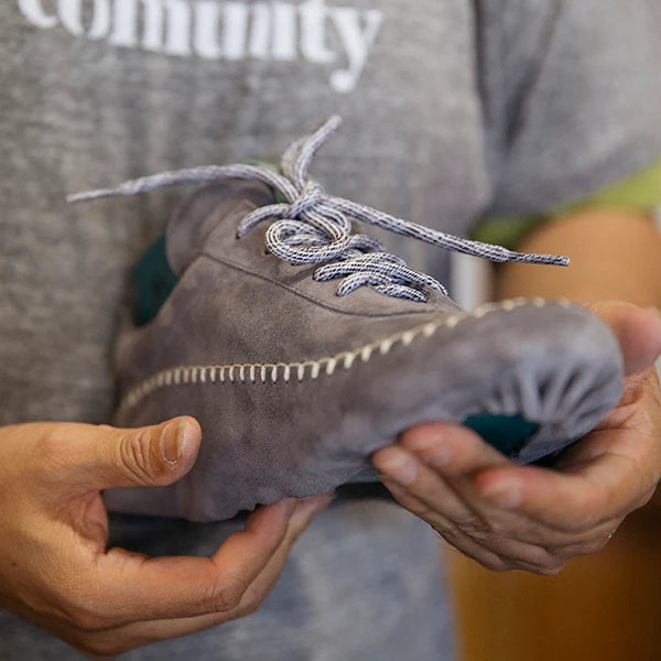 A close-up shot of a person holding a single, handmade grey suede shoe. The shoe features prominent white stitching along the top edge of the sole and on the toe. The laces are a speckled grey and white, and a pop of teal color is visible on the heel and inside the shoe. The person holding the shoe is wearing a grey t-shirt with the word "comunity" visible at the top of the frame.
