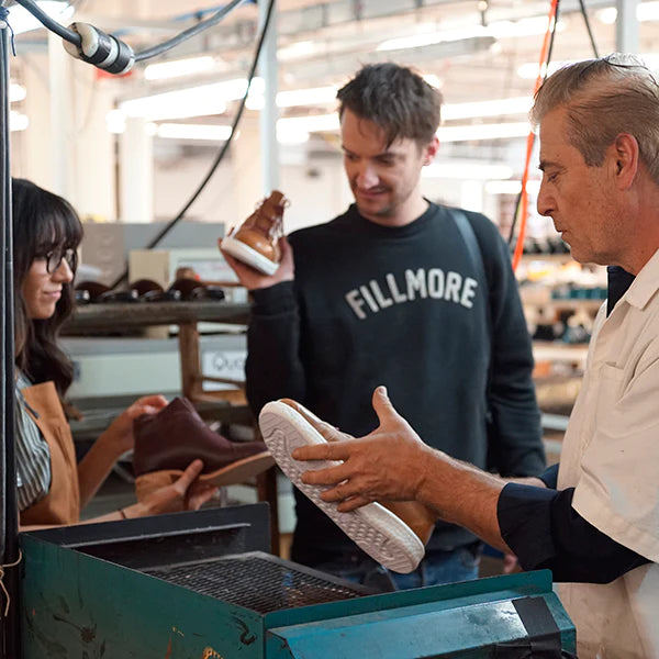 
A group of three people in what appears to be a shoe factory or workshop. A man on the right, wearing a white smock, holds a shoe with a white sole, showing it to a man in the center. The man in the center is wearing a black sweatshirt with the word "FILLMORE" on it and is holding a finished brown leather shoe. 