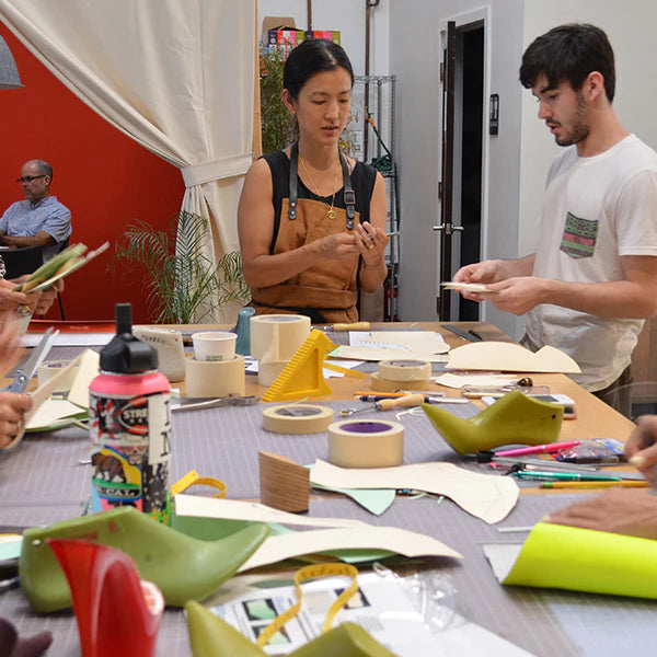 A woman and a man are working at a large table filled with shoe-making materials and tools. The woman, wearing a black top and a brown apron, is standing and looking down at a small object in her hands. The man next to her is holding a piece of paper or pattern. The table is cluttered with various objects including a water bottle with a colorful sticker, tape, rulers, and multiple green plastic shoe lasts. In the background, another person is sitting at a desk.