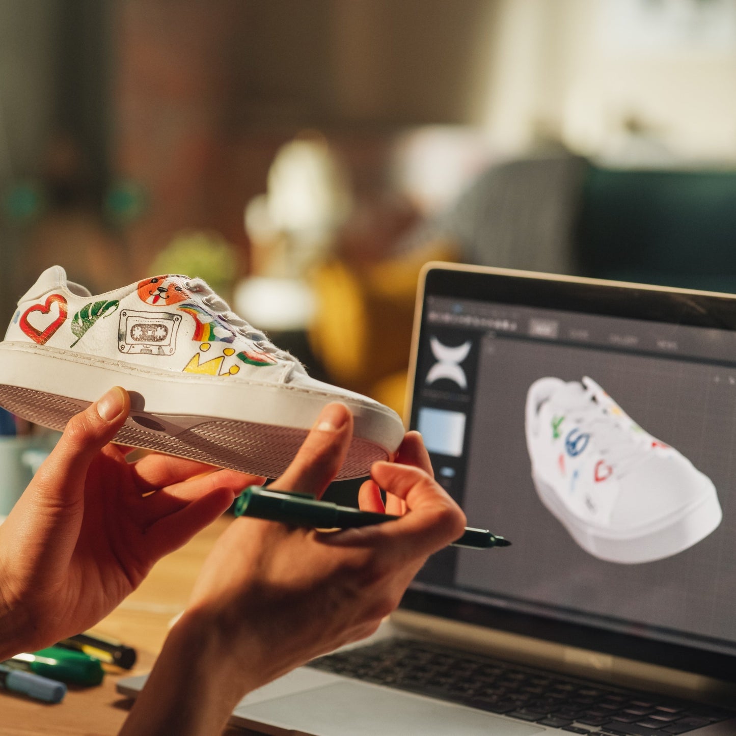 Photo of a person holding a white shoe that has drawings of a heart, crown, leaf, cat face on the shoe with a picture of a computer design of the shoe in the background