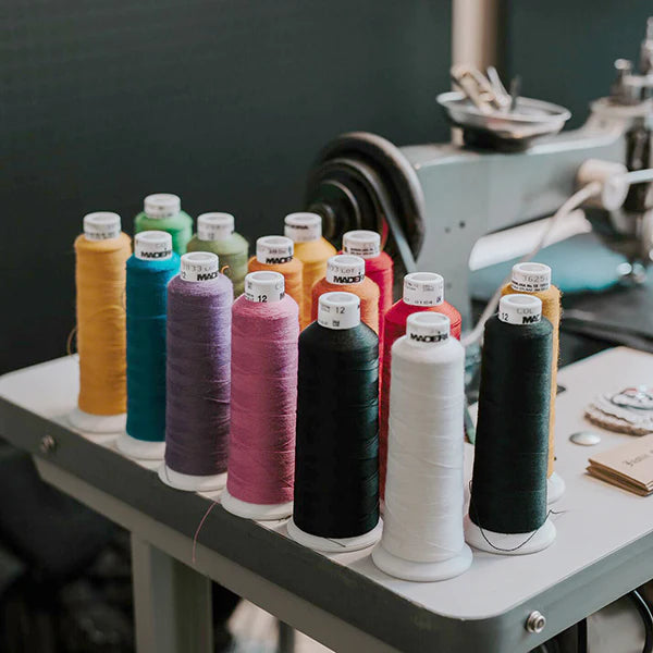 A close-up shot of a table with several large spools of thread in various colors, standing upright. The colors include mustard yellow, teal, light green, brown, deep purple, black, red, and white. In the blurry background, a sewing machine is visible, suggesting a crafting or workshop setting. The thread spools are organized in two rows.