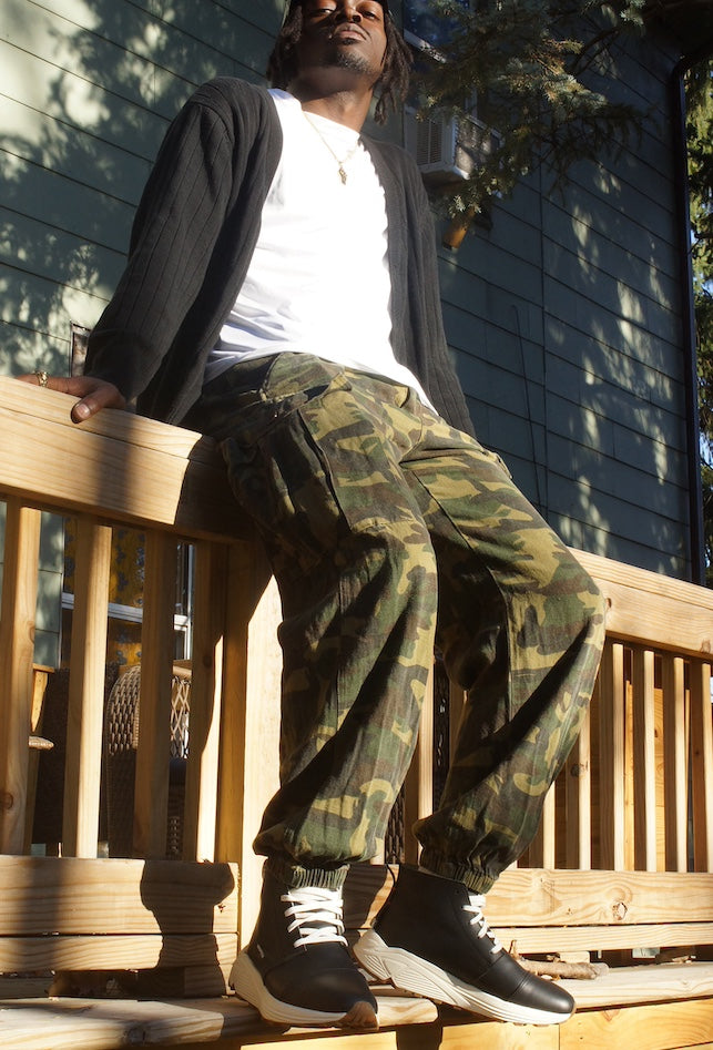 Photo of a man sitting on a fence with camo pants, white shirt and a grey sweater and black mid-cut sneakers that lace up with a white midsole and gum rubber outsole
