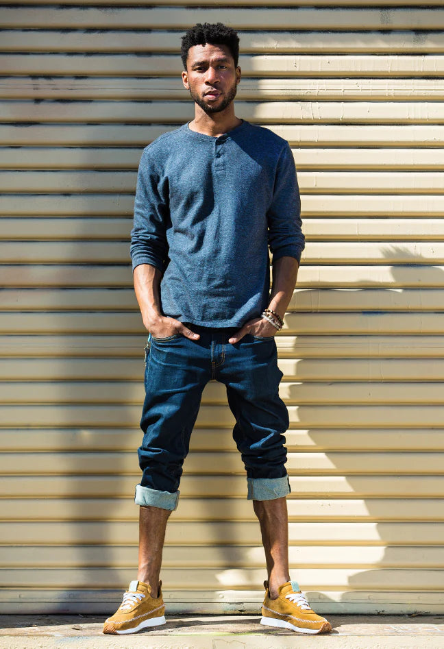 Front shot of a man wearing a blue shirt and blue jeans wearing brown suede sneakers that have stitching accents and a white outsole