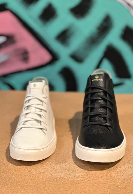 Photo of a half pair of white and half pair of black leather mid-cut sneakers shot from the front. They both have white rubber outsoles.