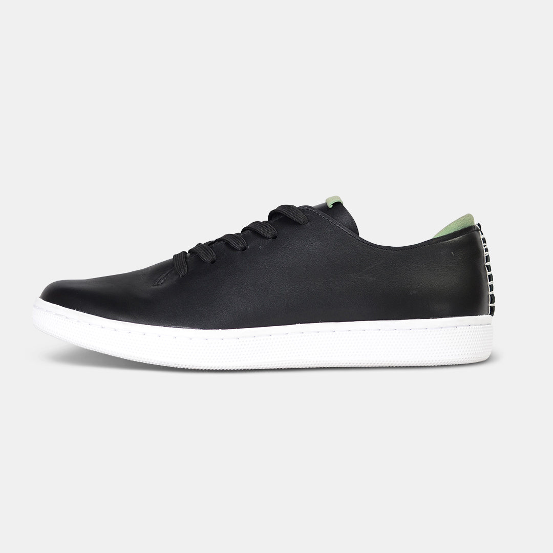 A profile shot of single black leather sneaker with a lace-up closure, featuring flat black shoelaces, a white rubber cupsole and a green accent on the heel.