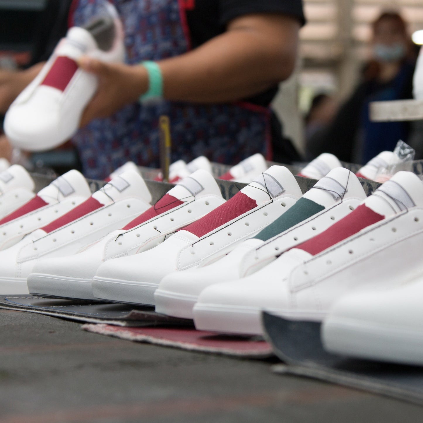 Photo of several pairs of white shoes without laces and patches of green and pink accents and a woman in the background holding a single shoe
