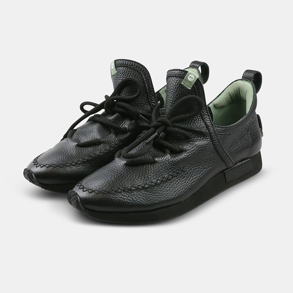 A quarter profile shot of a pair of casual black leather mid-cut slip-on shoe with unique stitch detailing on the toe and green branding accents.  Round black laces with heel loop pull tab and a black midsole and black rubber outsole.