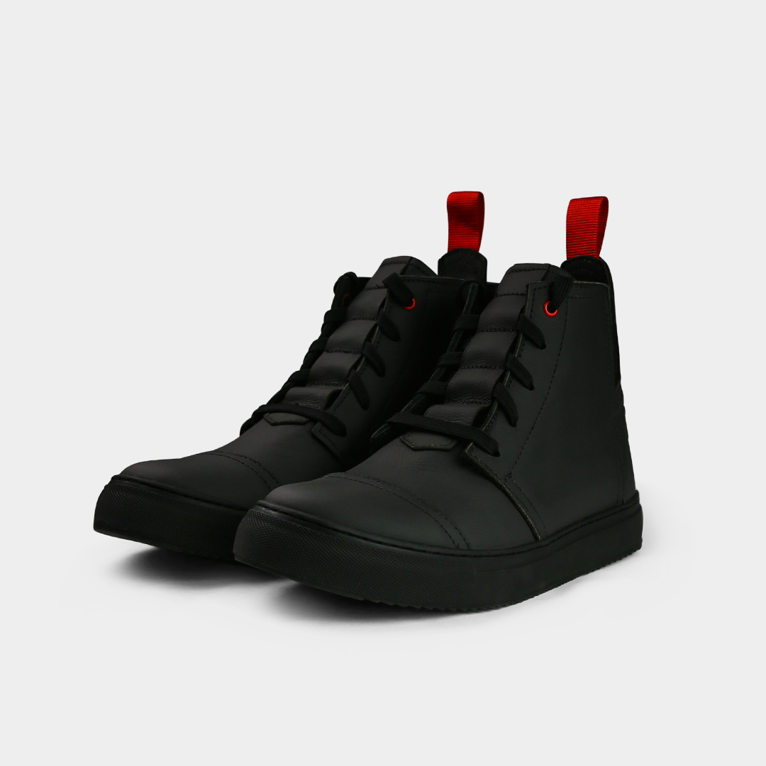 A low-angle product shot of a pair of black high-top shoes, placed side-by-side. The shoes are made of black leather, with black laces and a thick black sole. A bright red loop is attached to the back of each heel. The shoes have a sleek, modern design, and they are on a solid light gray background.