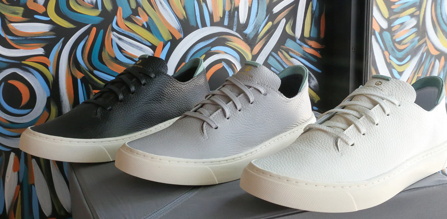 A product shot of three ComunityMade sneakers, lined up side-by-side, on a gray surface. The sneakers are a low-top style with white soles. From left to right, the shoes are black, light gray, and off-white. The material appears to be pebbled leather, and the heels have a pop of light green color. In the background, a colorful, abstract painting with swirling lines in blue, orange, and white is visible.