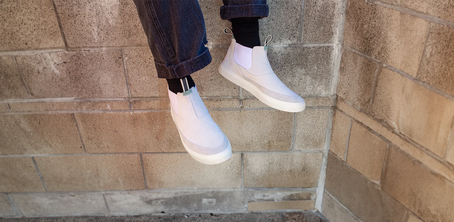 A pair of legs wearing black socks and dark, cuffed jeans is shown. A pair of off-white, high-top slip-on sneakers with white soles are on the feet. The Devium sneakers have a clean, minimalist design with a small green loop on the back of the ankle. The person is sitting on a ledge with their feet dangling in front of a brick wall.